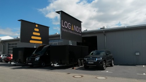 Two black vehicles are parked outside a gray industrial building with large LOGONDO Digital & Media Solutions Center signs, advertising LED trailer rentals. Shown is the LED Trailer 15 sqm—featuring a 5.12m x 2.88m V:LED VSF6 LED Screen—available for events and digital displays; the sky above is partly cloudy.