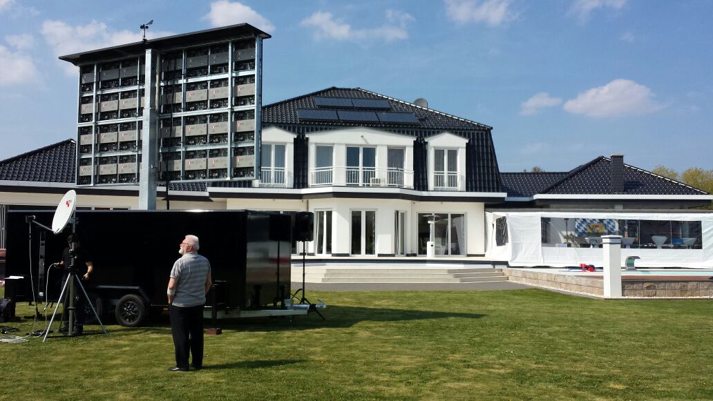 Modern white house with black roof tiles and large windows, featuring balconies. In the foreground, a man stands beside black equipment and a trailer. To the right, there is a tall fan structure and a white tent. Ideal SEO alt text for modern home exteriors or real estate listings.