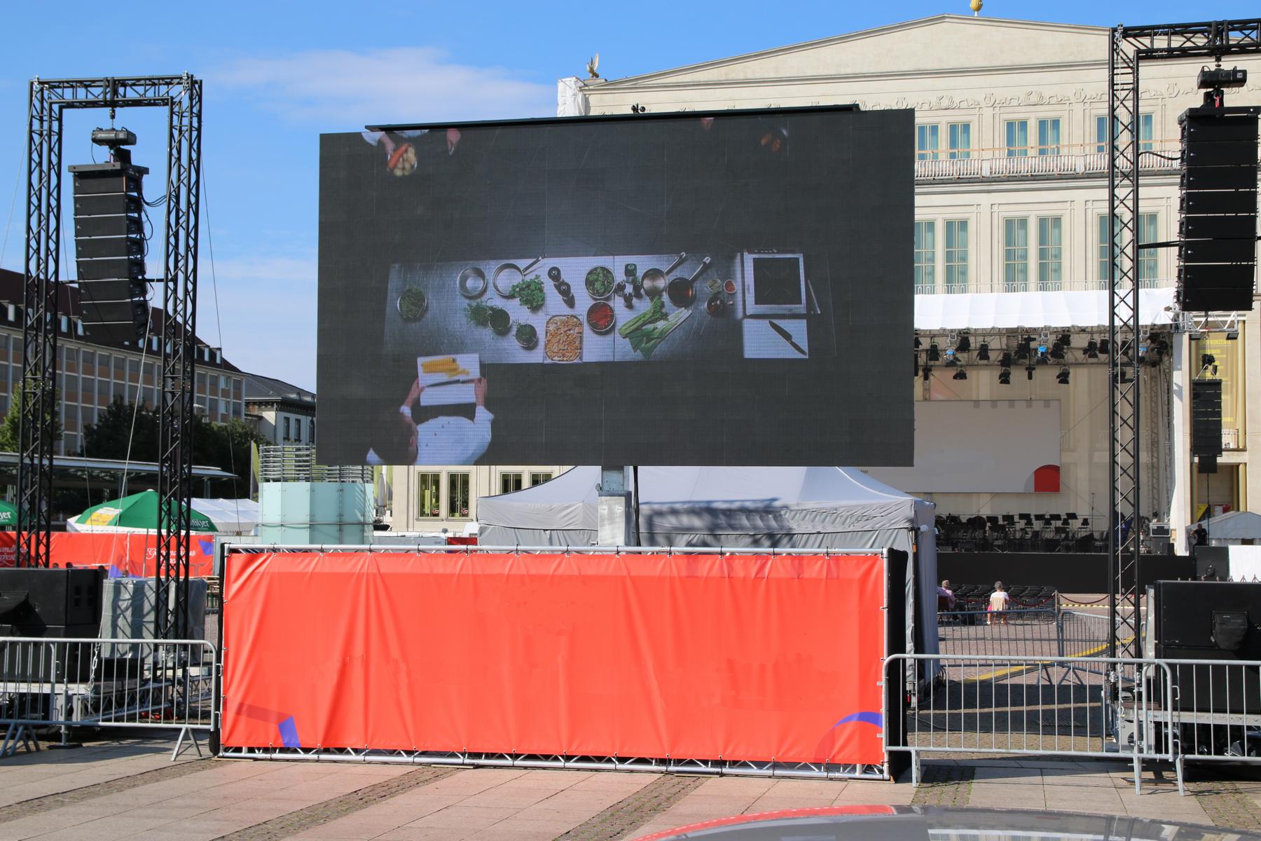 LED Trailer Cabrio 15 sqm (5.12m x 2.88m) V:LED VSF6 rental screen displayed outdoors in front of a grand columned building, showing a top-down image of food ingredients and a tablet; event barriers and white tents are set up nearby.
