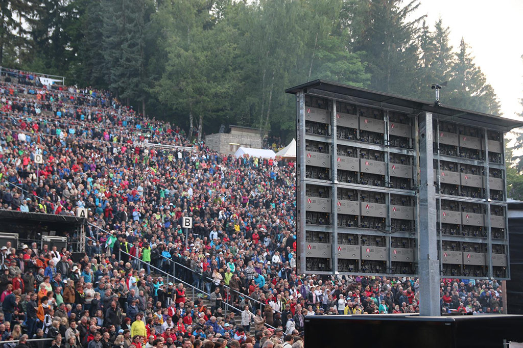 Large outdoor amphitheater with tiered seating filled with a crowd; tall trees surround the venue and a big screen or stage setup is visible in front as the audience watches an event. Perfect for open-air concerts and live performances.