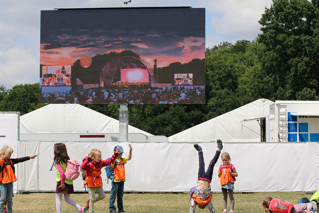 Children playing on grass at an outdoor event in front of a large concert screen at sunset, with kids wearing orange safety vests, white tents, and trees in the background. Family-friendly festival image.