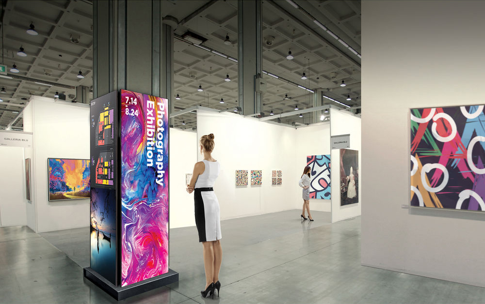 A woman in a stylish white and black dress stands in a bright, modern art gallery, admiring vibrant abstract paintings on the walls. In the center of the spacious room, a digital display advertises an upcoming photography exhibition.
