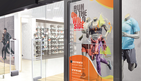 Storefront display at a sports shop featuring mannequins in colorful athletic wear behind a large window poster that reads RUN ON THE WILD SIDE. Shelves with athletic shoes and sporting goods are visible inside, showcasing running gear and fitness apparel.