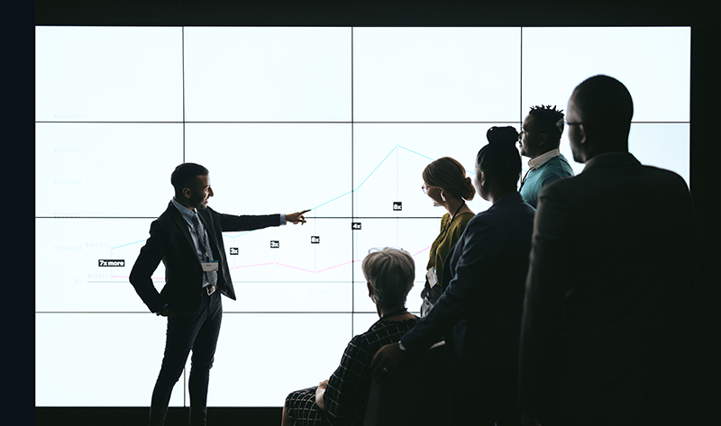 Business professional presenting data on a large screen with graphs to attentive colleagues in a dimly lit conference room. Perfect image for illustrating business meetings, corporate presentations, data analysis, or teamwork.