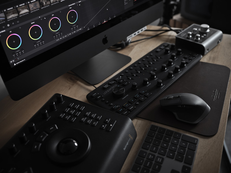 Desktop setup featuring a large monitor running color grading software, a specialized editing control panel, keyboard, mouse on a leather mousepad, and an audio interface arranged neatly on a wooden desk—ideal for professional video editing workstations.