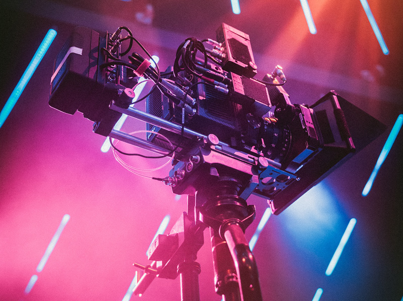 Professional video camera on tripod surrounded by vibrant pink and blue stage lights, with attached cables and equipment, creating a dramatic filming atmosphere – ideal for studio or live event production.
