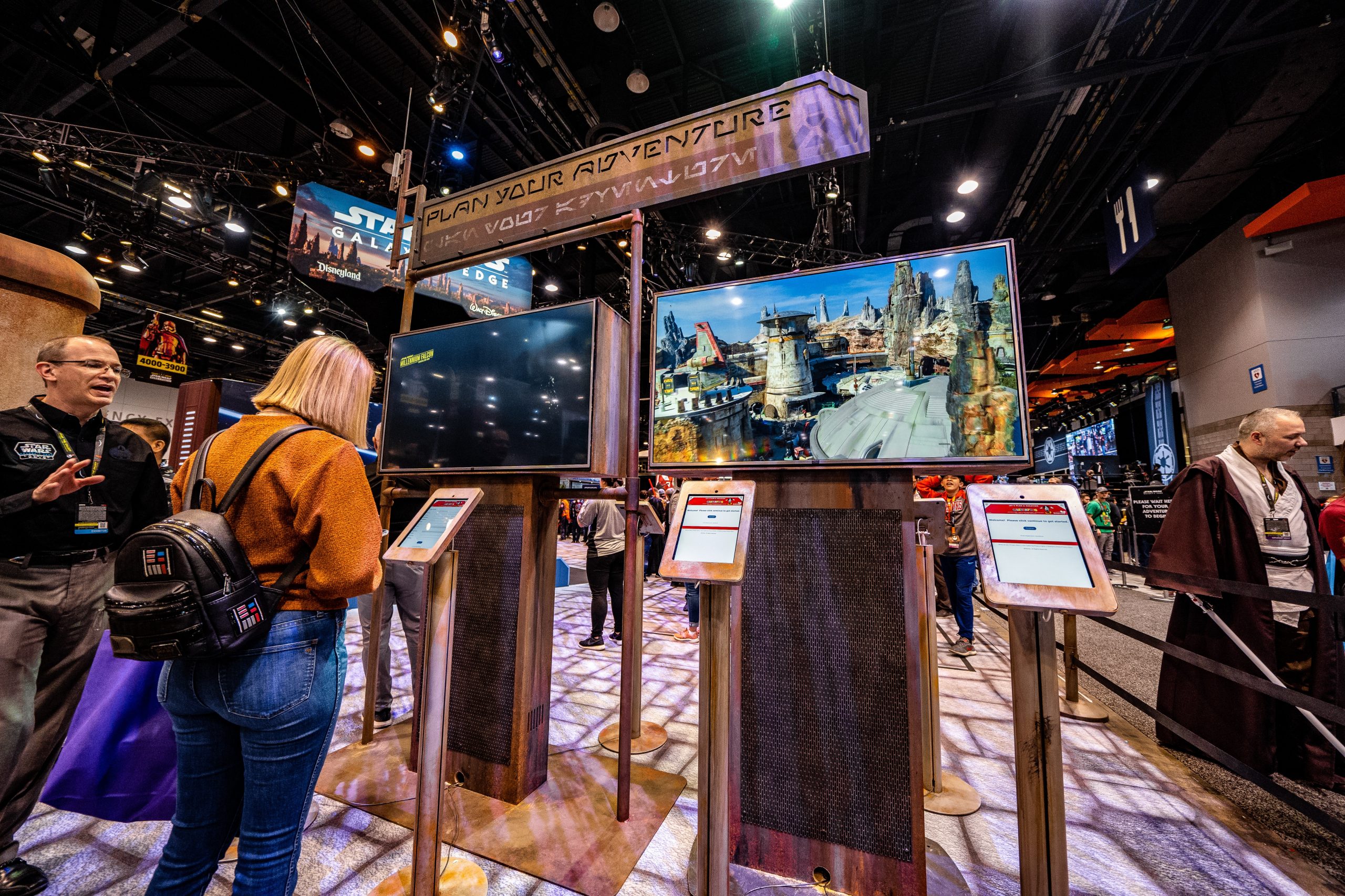 Woman using touchscreen kiosk at Star Wars event booth, with backpack, large screens showing sci-fi cityscape, and Plan Your Adventure sign above. Other attendees nearby.
