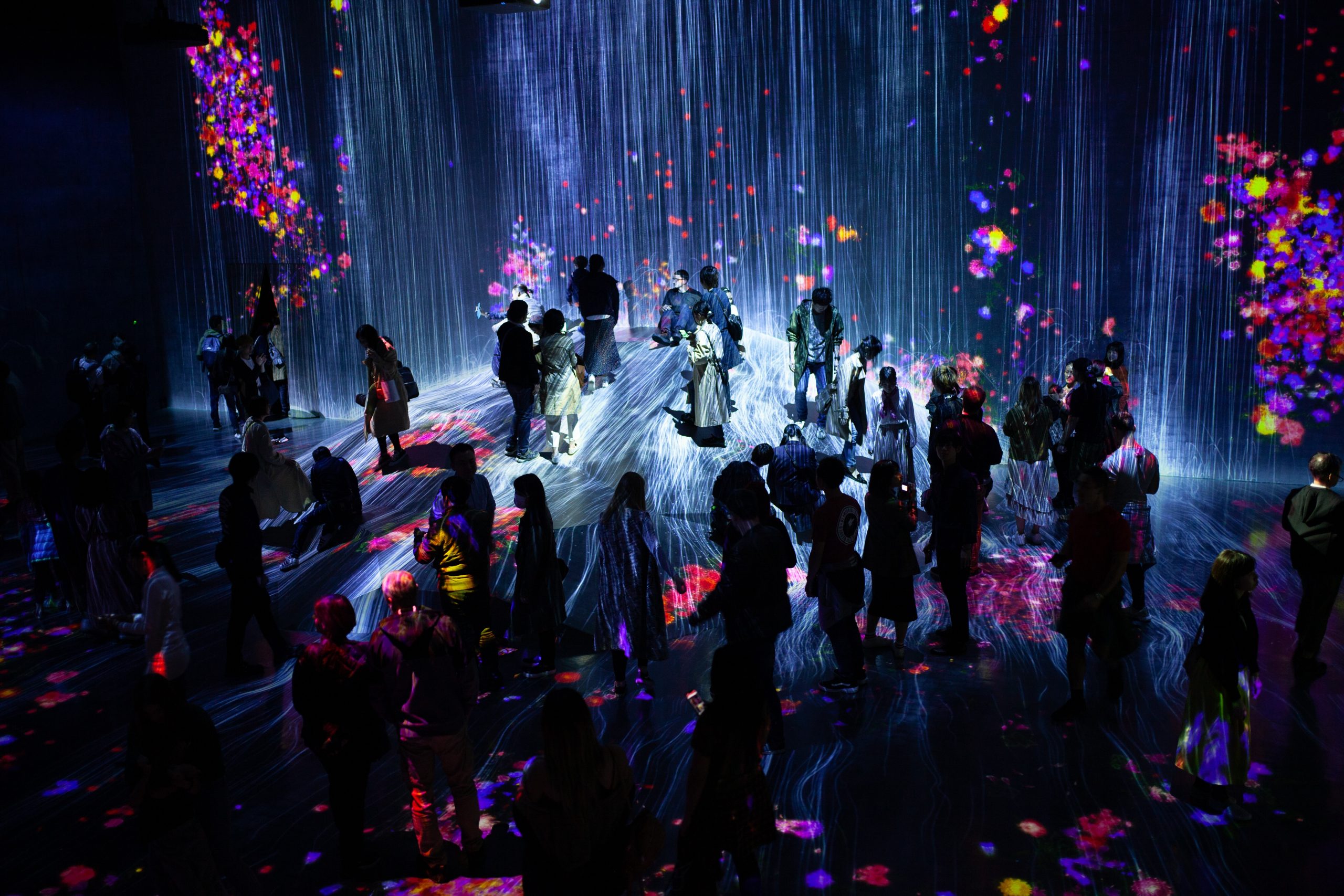 Visitors explore a dark room at the teamLab Borderless digital art museum, walking among vibrant, colorful lights and animated digital flower patterns projected onto the floor and walls, creating an immersive interactive art installation experience.