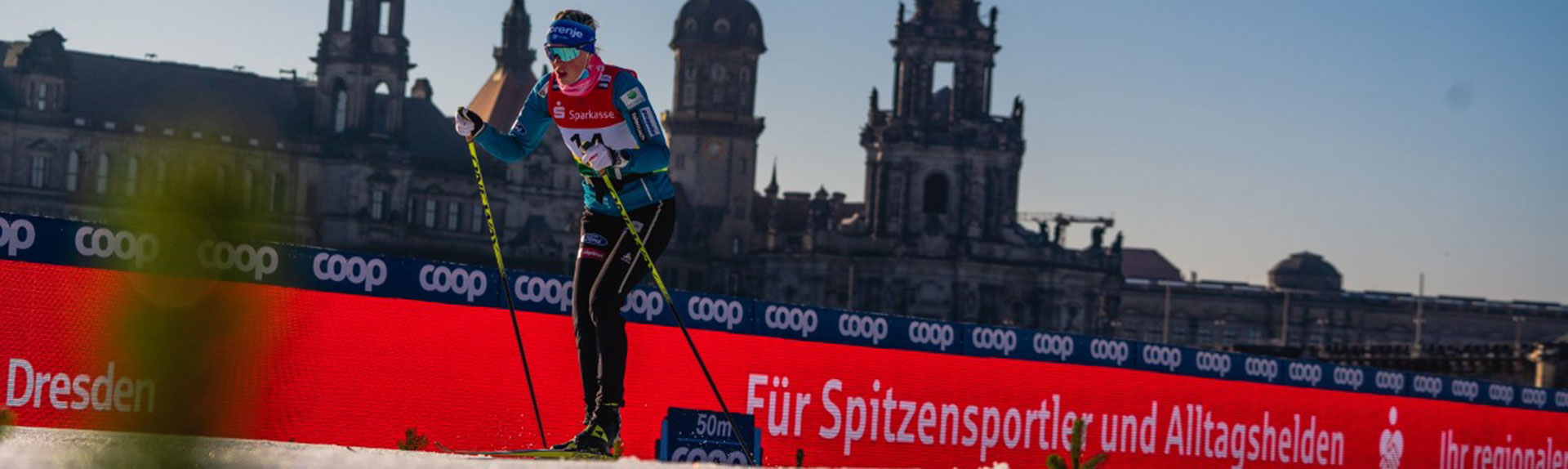A cross-country skier in racing gear glides along a snowy track with ski poles, passing a red advertising banner and historic buildings. A bright LED videowand rent display stands prominently beside the course under a clear blue sky, creating an eye-catching backdrop for the winter sports event.