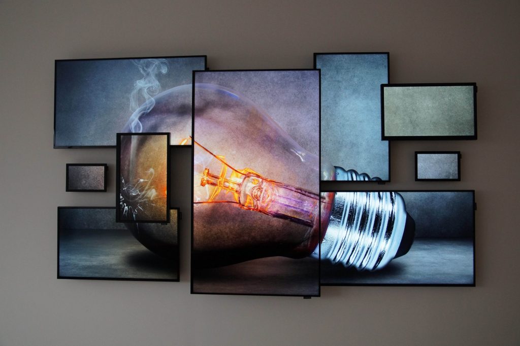 Digital art installation in a modern office showing a large light bulb displayed across multiple screens of different sizes, creating a fragmented but unified image against a plain background. Ideal for innovative workspace décor and contemporary digital office design.