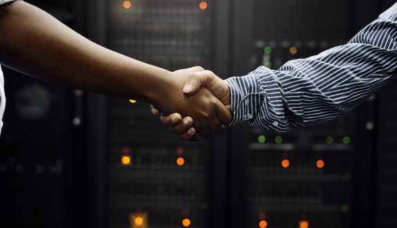 Two people shaking hands in front of illuminated computer servers, symbolizing partnership and collaboration in technology for the Logando About Us page.