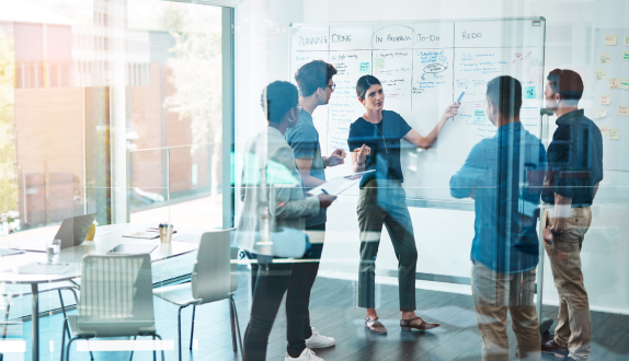 Five team members in a modern office collaborate at a whiteboard filled with notes and diagrams, discussing Logando About Us and teamwork strategies; laptops and materials are visible, emphasizing team collaboration and workplace productivity.